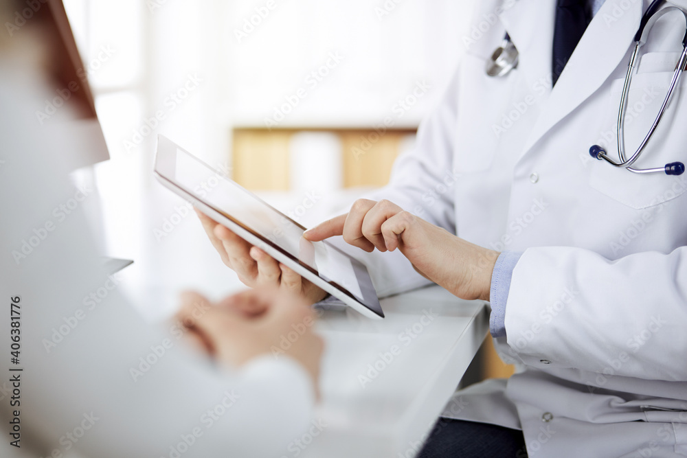 Unknown male doctor and patient woman discussing current health examination while sitting in sunny clinic and using tablet computer. Perfect medical service in hospital Unknown male doctor and patient woman discussing current health examination while sitting in sunny clinic and using tablet computer. Perfect medical service in hospital