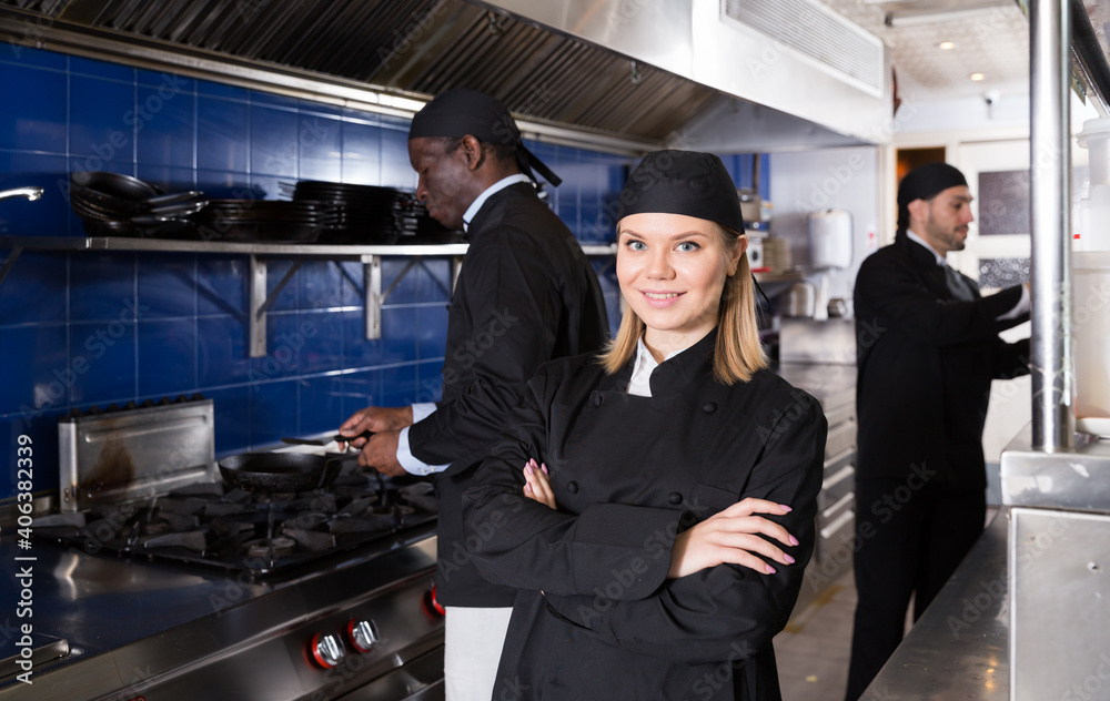 Portrait of confident young woman chef in modern restaurant kitchen with working staff