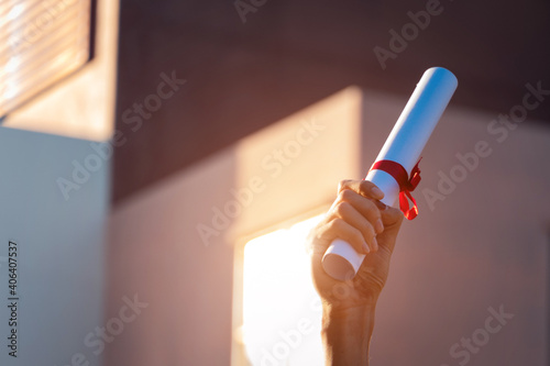 Close-up shot of a university graduate holding degree certification to shows and celebrate his success in the college commencement day with sunlight in the background.