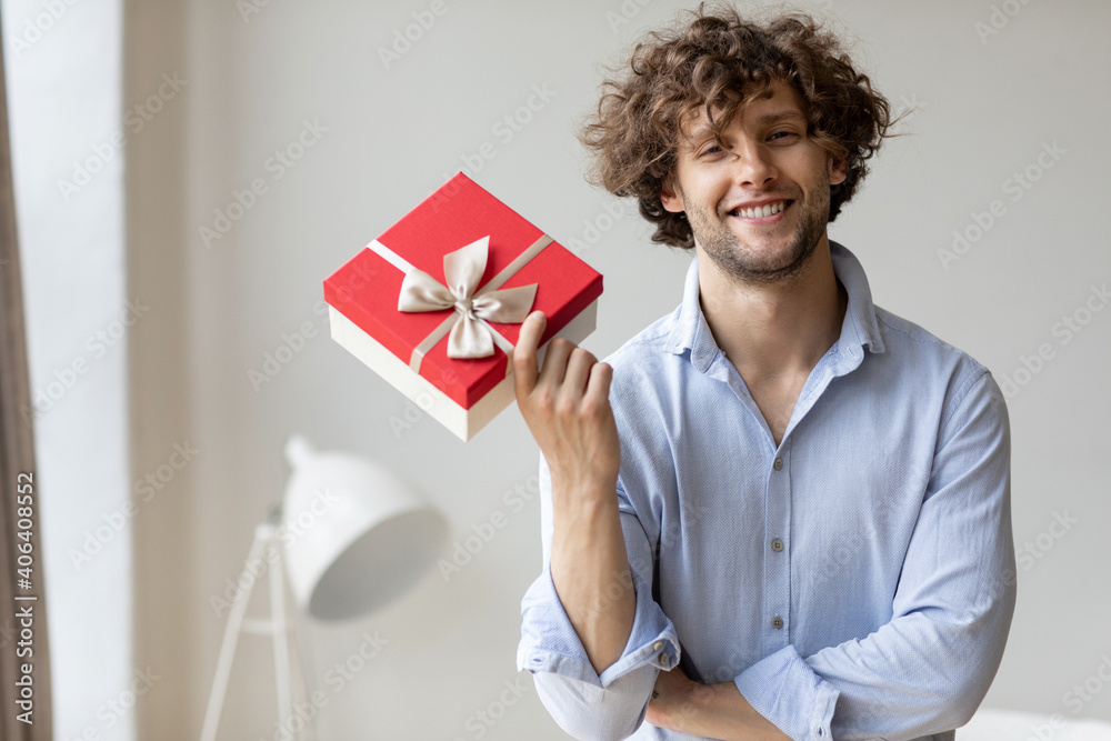 Handsome man holding a gift in a blue shirt with curly hair at home.