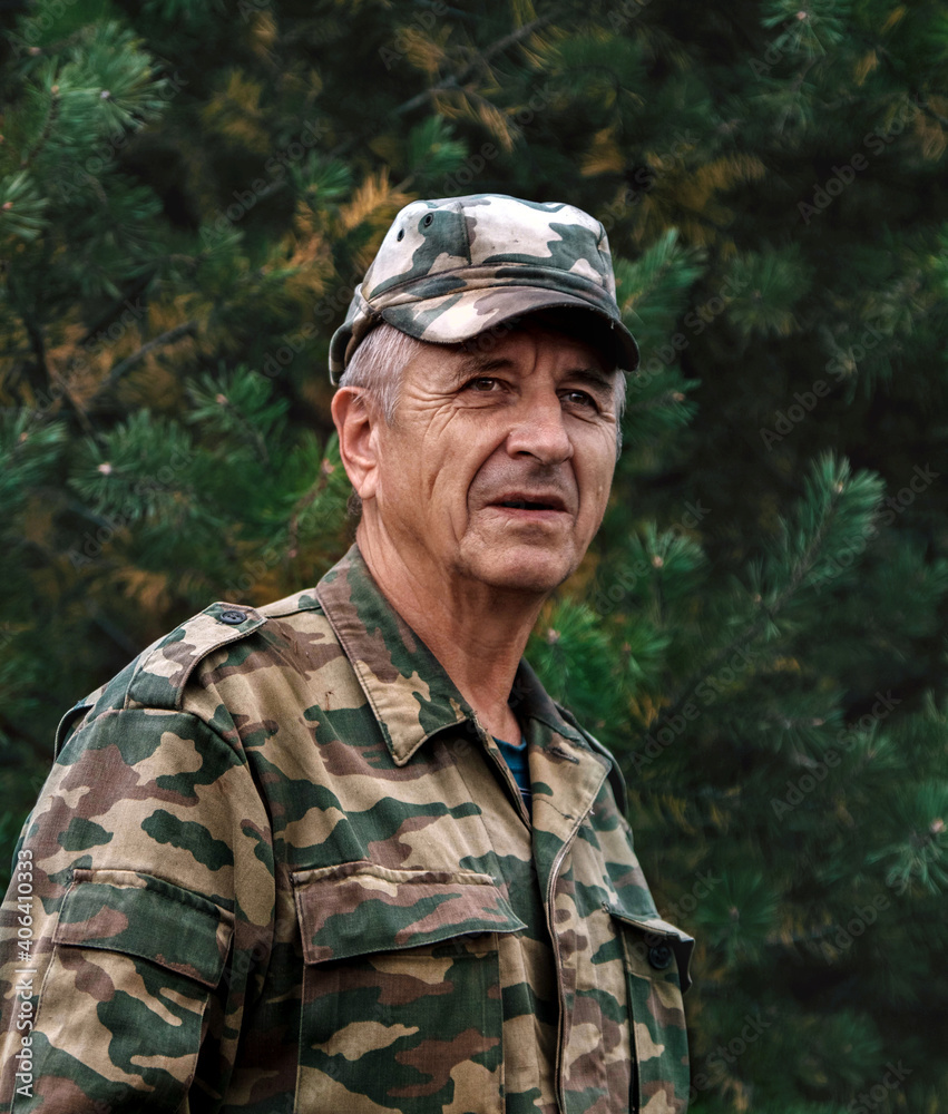 Elderly man in motley jacket and kepi against green branches of pine tree