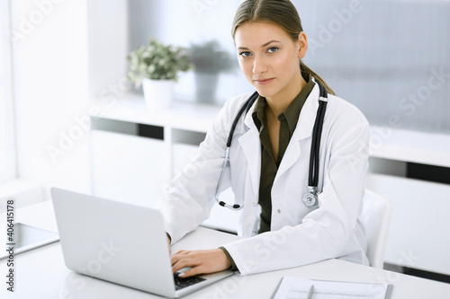 Woman-doctor typing on laptop computer while sitting at the desk in hospital office. Physician at work. Data in medicine and healthcare