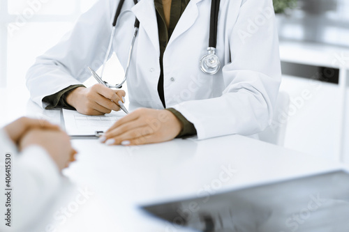 Unknown woman-doctor and female patient sitting and talking at medical examination in clinic, close-up. Therapist wearing green blouse is filling up medication history record. Medicine concept