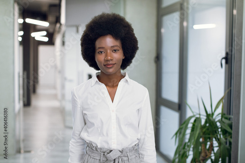 Half-length portrait of charming young purposeful African businesswoman with afro hair, wearing white shirt and gray pants, posing to camera indoors, in modern loft office. Successful lady