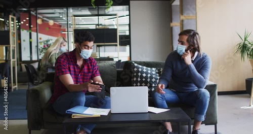 Diverse business people wearing masks using laptop and goign through paperwork in modern office photo