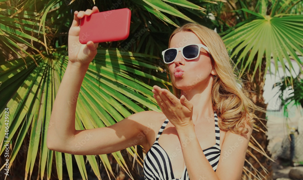 Summer portrait of young woman taking selfie picture by smartphone blowing lips sending sweet air kiss on a beach, palm tree background Summer portrait of young woman taking selfie picture by smartphone blowing lips sending sweet air kiss on a beach, palm tree background
