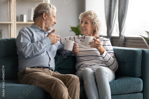 Bonding elderly family couple drinking morning coffee or hot tea, enjoying pleasant conversation relaxing together on cozy sofa in living room, retired people lifestyle, good relations concept.