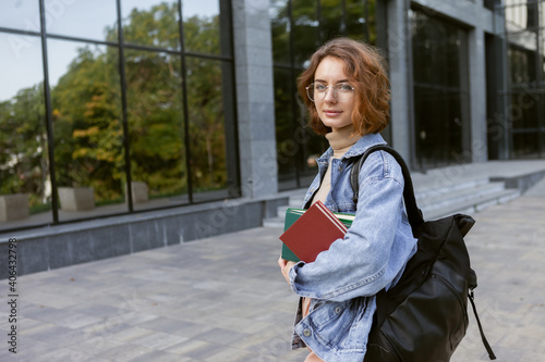 Modern attractive woman student in stylish clothes with stack of books and backpack outdoors