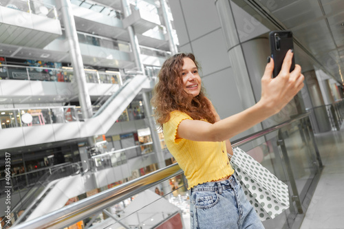 Cute woman with curly hair takes a selfie with shopping bags in a shopping center. Shopaholic concept