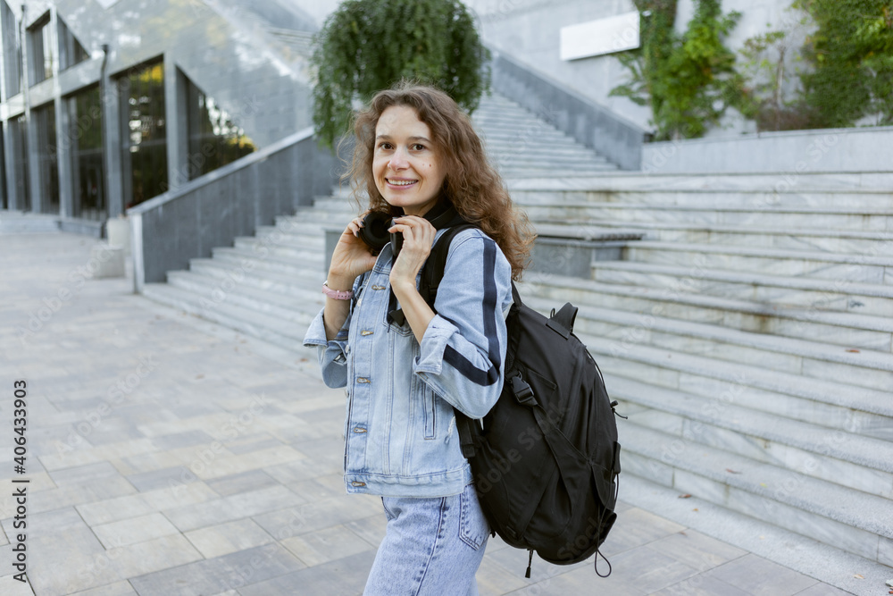 Portet charismatic curly hair woman in denim jacket with backpack and headphones outdoors in city