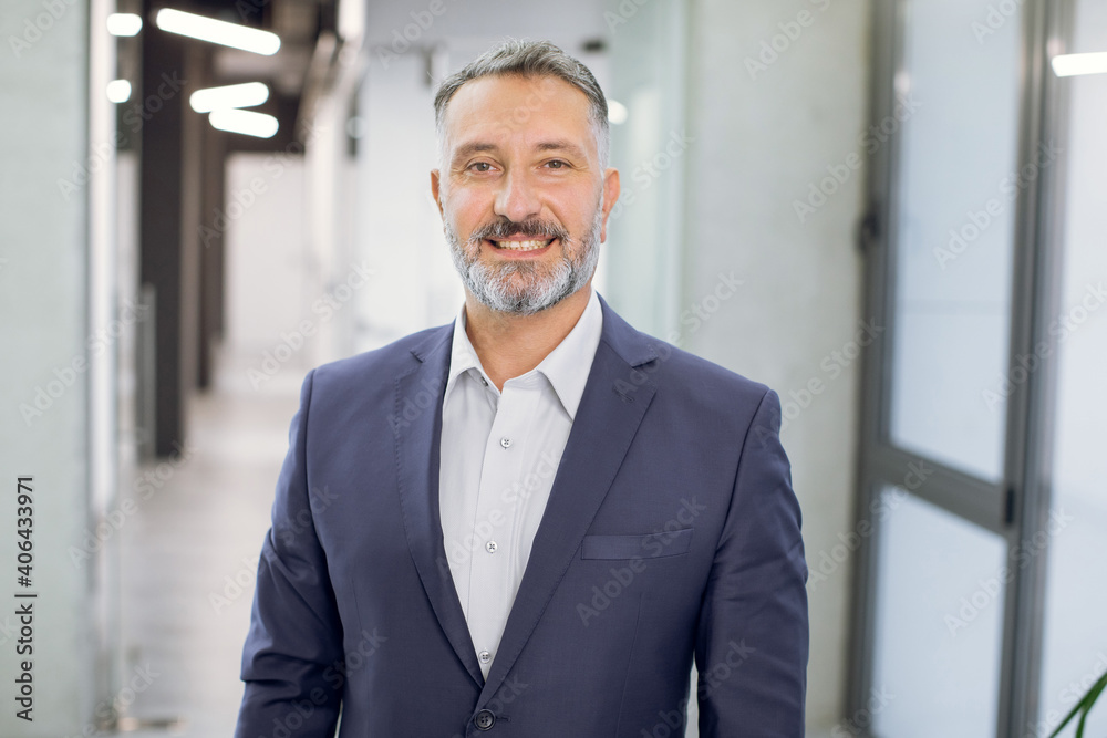 Close up front view of happy 50-aged bearded businessman in formal wear, posing to camera with smile, while standing in modern light office corridor. Indoor portrait of mature office worker