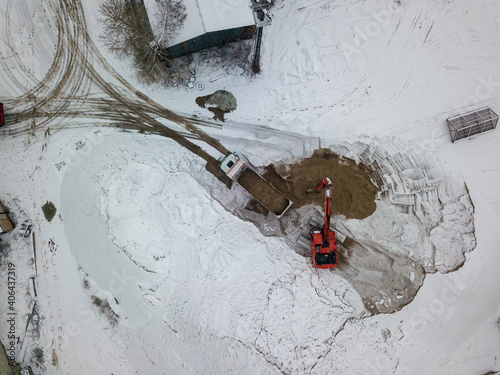 An excavator loads sand into a truck. Snowy day, blizzard. Aerial drone top view.