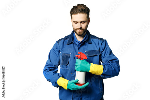 Young handsome man with a beard in a blue working uniform for cleaning rooms in rubber gloves and a janitor's uniform holds chemical spray bottle for cleaning. Isolated on a white background