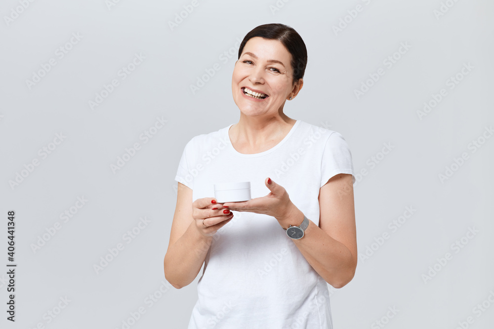 Pretty middle aged woman is smiling and looking at camera with anti age cream. Isolated on gray background. Pretty middle aged woman is smiling and looking at camera with anti age cream. Isolated on gray background.