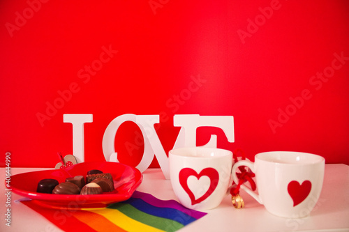 red plate in the shape of a heart with bonbons and two cups white with red hearts and gay pride flag