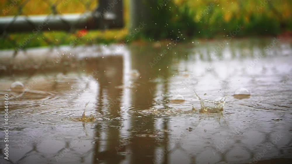 Raindrops falling from the sky and splashing in a backyard puddle by a chain-link fence or gate - slow motion Raindrops falling from the sky and splashing in a backyard puddle by a chain-link fence or gate - slow motion