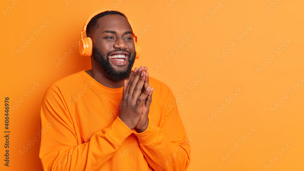 Happy plump dark skinned man keeps palms pressed together smiles broadly dressed in casual sweater wears stereo headphones listens pleasant melody poses against orange studio wall with copy space Happy plump dark skinned man keeps palms pressed together smiles broadly dressed in casual sweater wears stereo headphones listens pleasant melody poses against orange studio wall with copy space