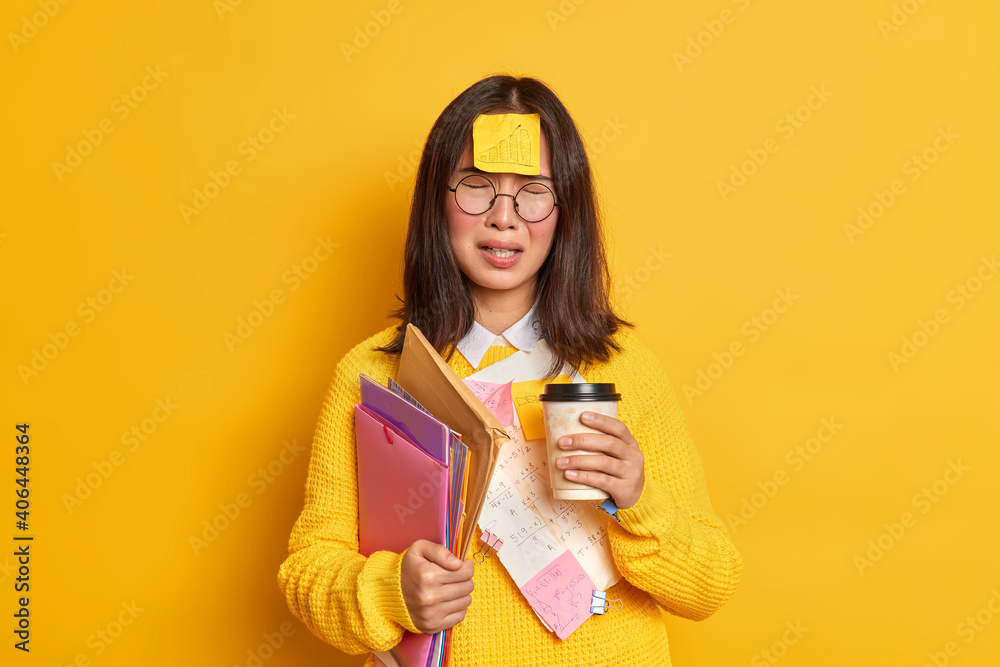 People and education concept. Upset Asian female student feels tired of exam preparation drinks takeaway coffee makes memo stickers has bad mood cannot remember everything holds folders with papers People and education concept. Upset Asian female student feels tired of exam preparation drinks takeaway coffee makes memo stickers has bad mood cannot remember everything holds folders with papers