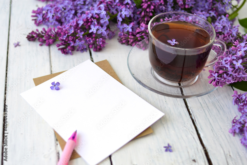 Delicate morning tea table setting with lilac flowers, a transparent cup of tea and a saucer and a white vase, white and craft paper for notes, and a pink pen on a white wooden table. Mockup. Delicate morning tea table setting with lilac flowers, a transparent cup of tea and a saucer and a white vase, white and craft paper for notes, and a pink pen on a white wooden table. Mockup.