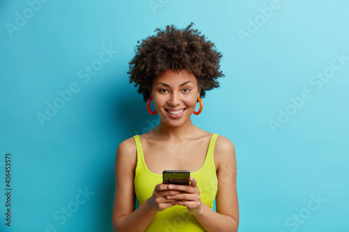 Glad African American woman uses mobile phone for checking message smiles positively wears casual clothes has curly bushy hair poses against blue background. Online lifestyle technology concept