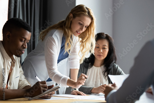 Serious millennial blonde female team leader analyzing financial or marketing paper document, reporting research results to focused multiracial young and older business partners colleagues at office.