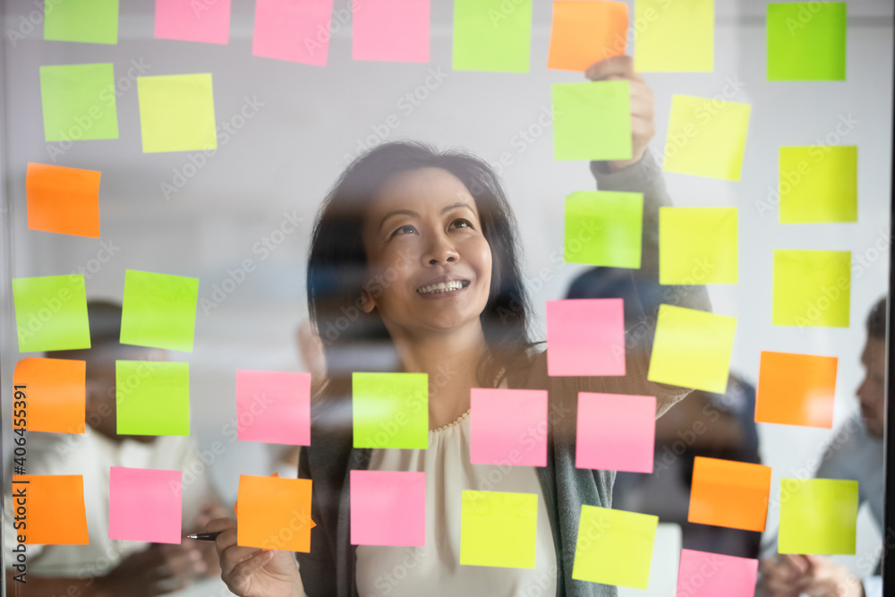 Happy middle aged korean female project manager standing behind glass wall, adding colored sticky notes on window, organizing working processes or adding steps, developing corporate strategy. Happy middle aged korean female project manager standing behind glass wall, adding colored sticky notes on window, organizing working processes or adding steps, developing corporate strategy.