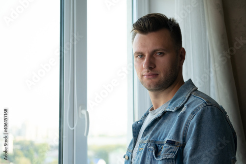 A handsome stylish European man in a jeans jacket stands at the window and looks at the camera