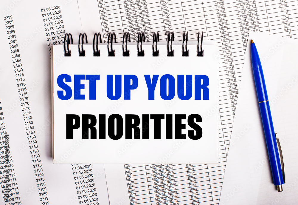 On the table are charts and reports, on which lie a blue pen and a notebook with the word SET UP YOUR PRIORITIES On the table are charts and reports, on which lie a blue pen and a notebook with the word SET UP YOUR PRIORITIES