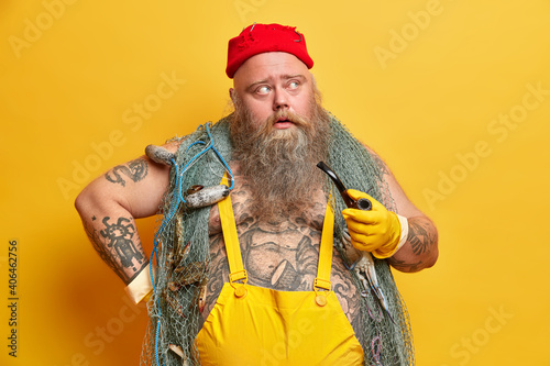 Thoughtful concentrated man seafarer with thick beard smokes pipe thinks about something dressed in overalls poses against yellow background. Pensive tattooed sailor has sea cruise or adventure