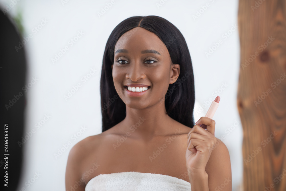 Smiling millennial african american lady with perfect skin looks in mirror and holds nude lipstick Smiling millennial african american lady with perfect skin looks in mirror and holds nude lipstick