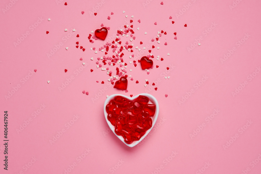 White ceramic heart shaped bowl with splash of red heart shaped confetti and small decorative hearts over pink background. Valentines Day concept. Top view, copy space.