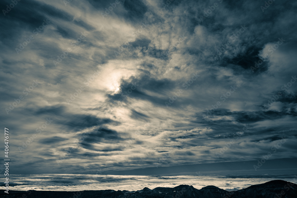 Cloudscape of high clouds of various types over mountains. Sun is barely visible from the clouds in the centre. The lower level of clouds in the background behind the black mountain ridge.
