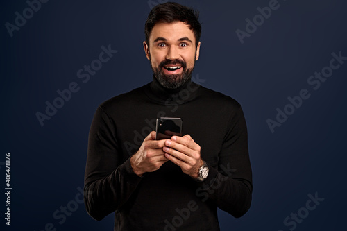 Cheerful, smiling guy, stylish hairstyle in high neck sweater, holding smartphone, look camera pleased and fascinated, receive great news via message in app, ordered product with great discount
