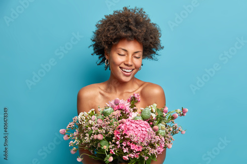 Spring is coming. Cheerful Afro American woman stands bare shoulders with closed eyes holds beautiful bouquet of flowers over naked body has date with boyfriend isolated over blue background
