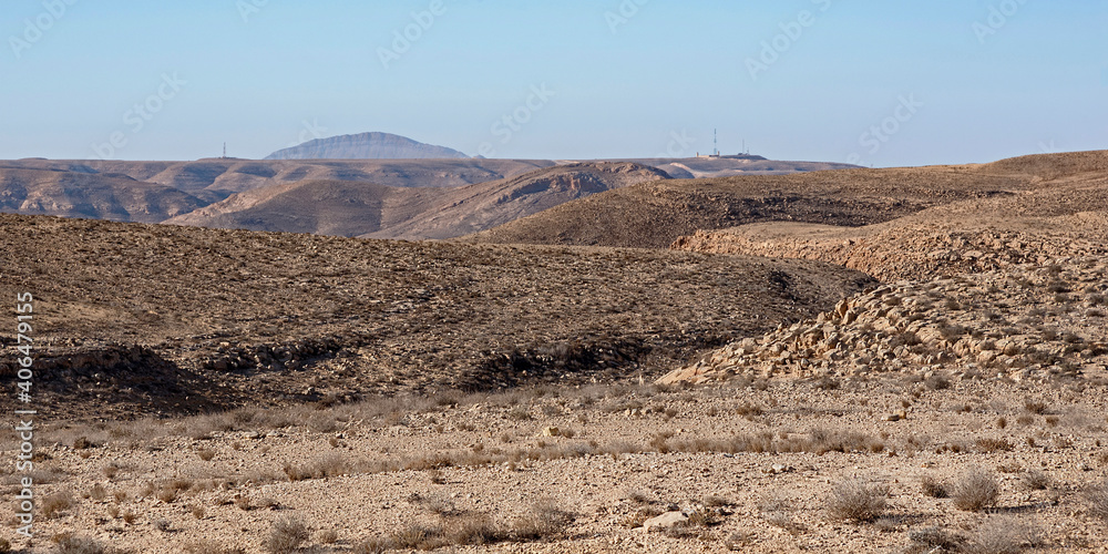 a rugged barren mountainous desert landscape west of the makhtesh ramon crater with an israeli border outpost and camel hump mountain in the sinai in the background against a clear blue sky