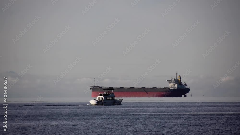 Giant empty Bulk carrier with dropped anchor waiting for orders while a small white yacht is passing on the foreground on a cloudy day. Steady wide shot Giant empty Bulk carrier with dropped anchor waiting for orders while a small white yacht is passing on the foreground on a cloudy day. Steady wide shot
