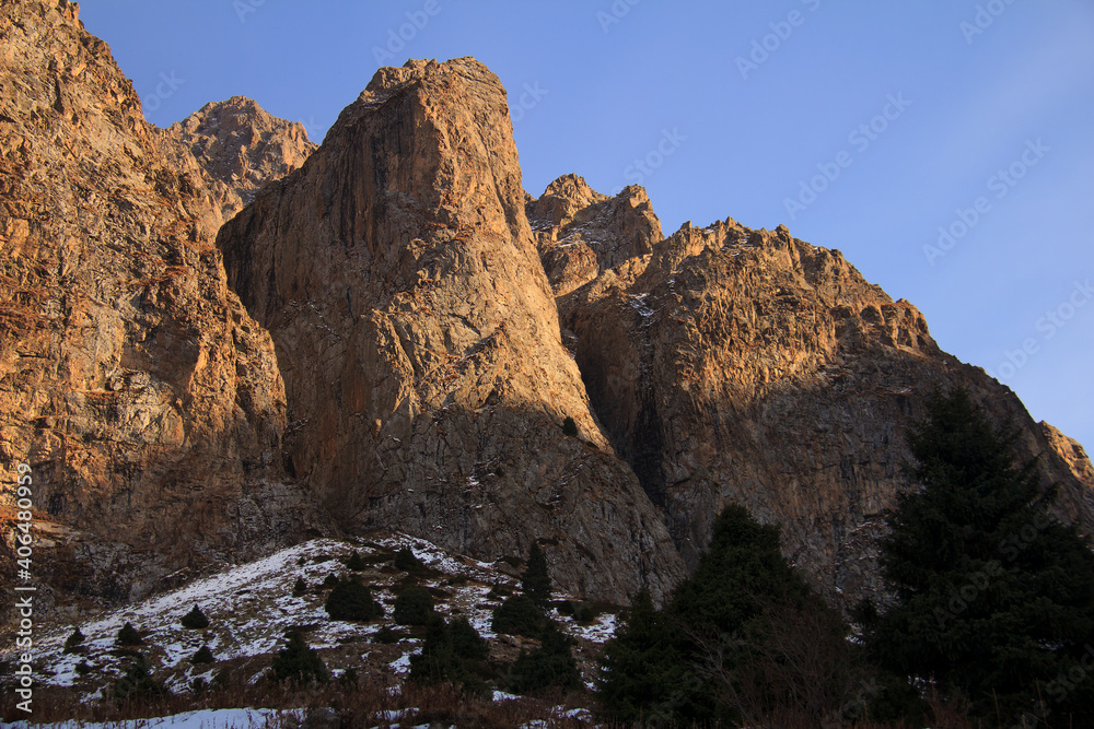 Huge rocky mountains with snow and trees at sunset in autumn. Large sheer cliffs against the background of a clear sky, below there is snow and trees grow, autumn, sunny
