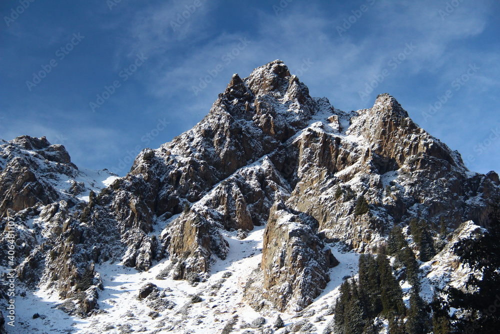 Peaked alpine cliffs covered with snow, clear day. Rocky alpine peak in the snow against a blue sky with small clouds, trees grow on the right, morning, sunny weather, in winter