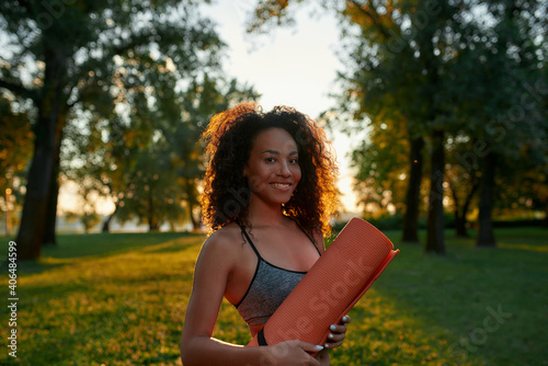 Young beautiful mixed race fitness woman wearing sport clothes holding yoga mat, smiling at camera while standing in the green park