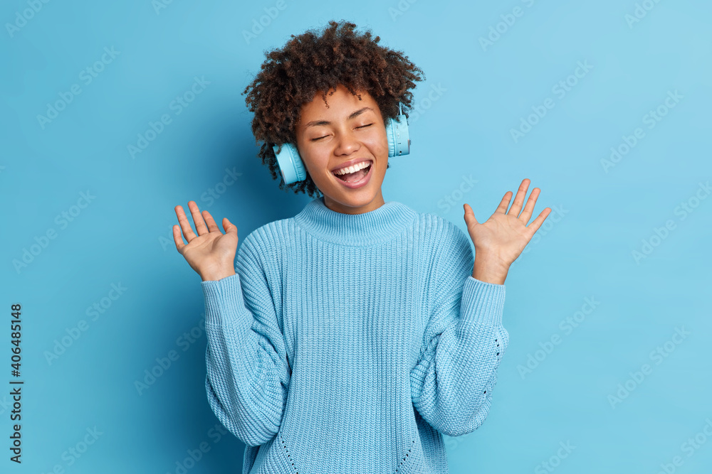 Positive African American woman with curly hair raises palms has fun while listening audio track wears wireless headphones dressed in casual jumper isolated over blue background. Favorite hobby