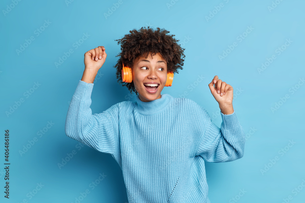 Waist up shot of joyful African American woman dances with rhythm of music wears stereo wireless headphones wears casual jumper isolated over blue background. Having fun. Happy female meloman Waist up shot of joyful African American woman dances with rhythm of music wears stereo wireless headphones wears casual jumper isolated over blue background. Having fun. Happy female meloman