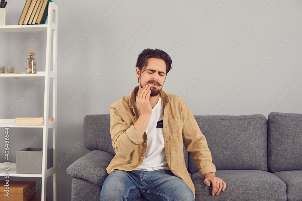 Dental problems concept. Upset young man suffering from acute toothache, inflamed nerve, tooth abscess, or teeth sensitivity. Sad male patient sitting on sofa and touching cheek with grimace of pain