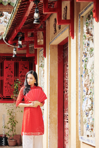 Beautiful pensive young Asian woman with red paper fan standing at temple doors and looking away © DragonImages
