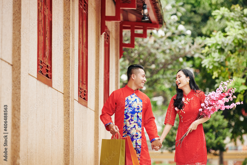 Obraz premium Happy young Asian couple holding hands when walking outdoors in traditional clothing with blooming branches and shopping bags