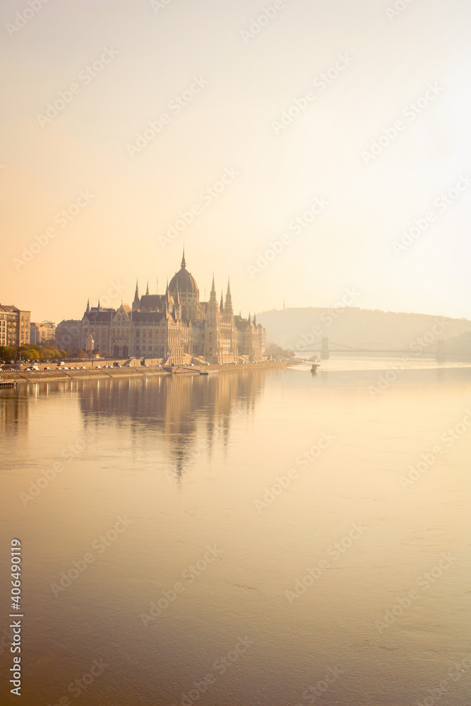 Hungarian Parliament landscape in sunset light next to the Danube with boats with foggy air and Gellert hill in the background and the Chain bridge