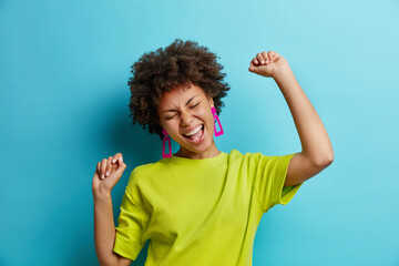 Overjoyed African American woman makes winning gesture expresses happiness raises arms and dances carefree celebrates victory dressed in t shirt isolated over blue background. Female triumphant