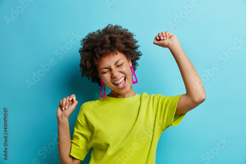 Overjoyed African American woman makes winning gesture expresses happiness raises arms and dances carefree celebrates victory dressed in t shirt isolated over blue background. Female triumphant