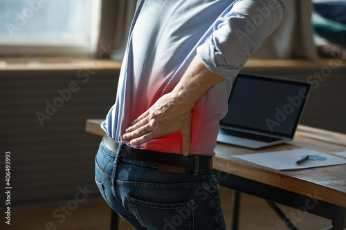 Close up rear view stressed young man touching lower back feeling discomfort, suffering from sudden pain due to sedentary lifestyle or long computer overwork in incorrect posture at home office.