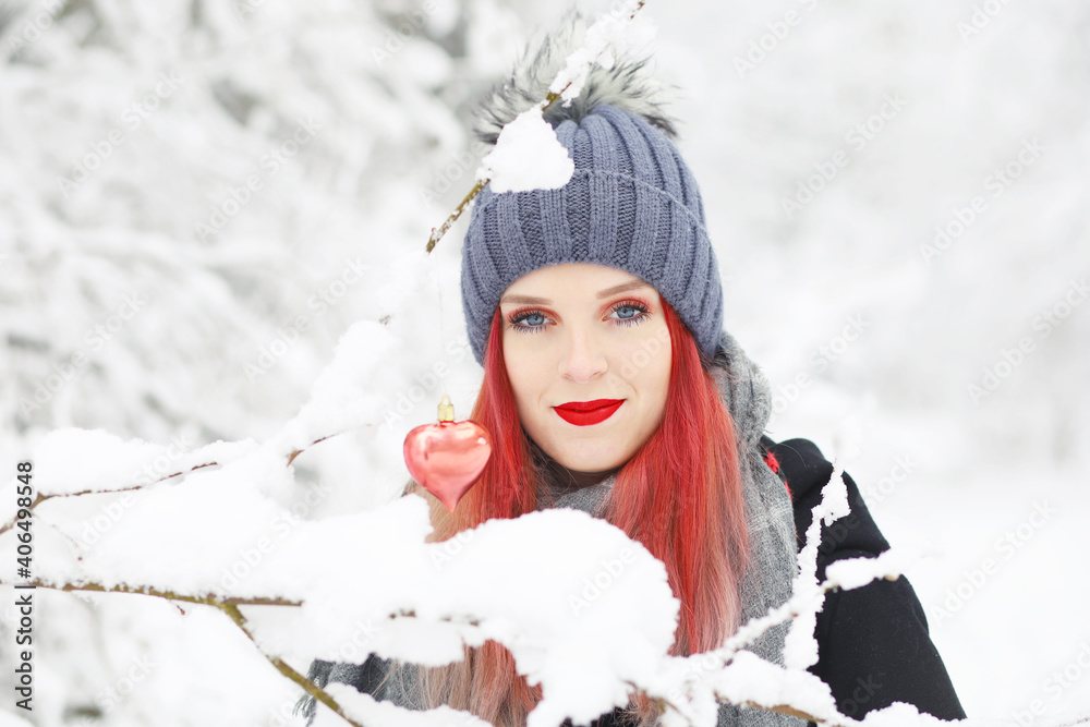Smiling attractive red hair girl is posing with red heart hanging on a frozen snowy branch. Smiling attractive red hair girl is posing with red heart hanging on a frozen snowy branch.
