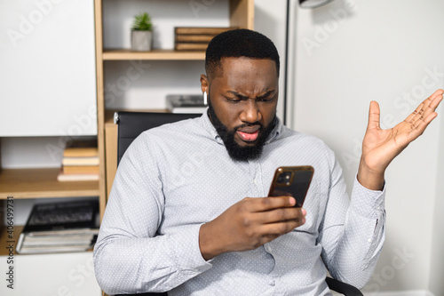 Confused puzzled African-American man in smart casual shirt holding smartphone and expressing misunderstanding, spreads his hands in incomprehension photo